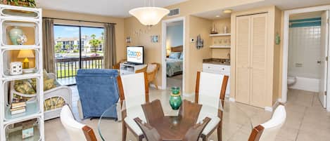 A modern living room with a glass dining table, a TV, and a balcony with a view of the neighborhood.