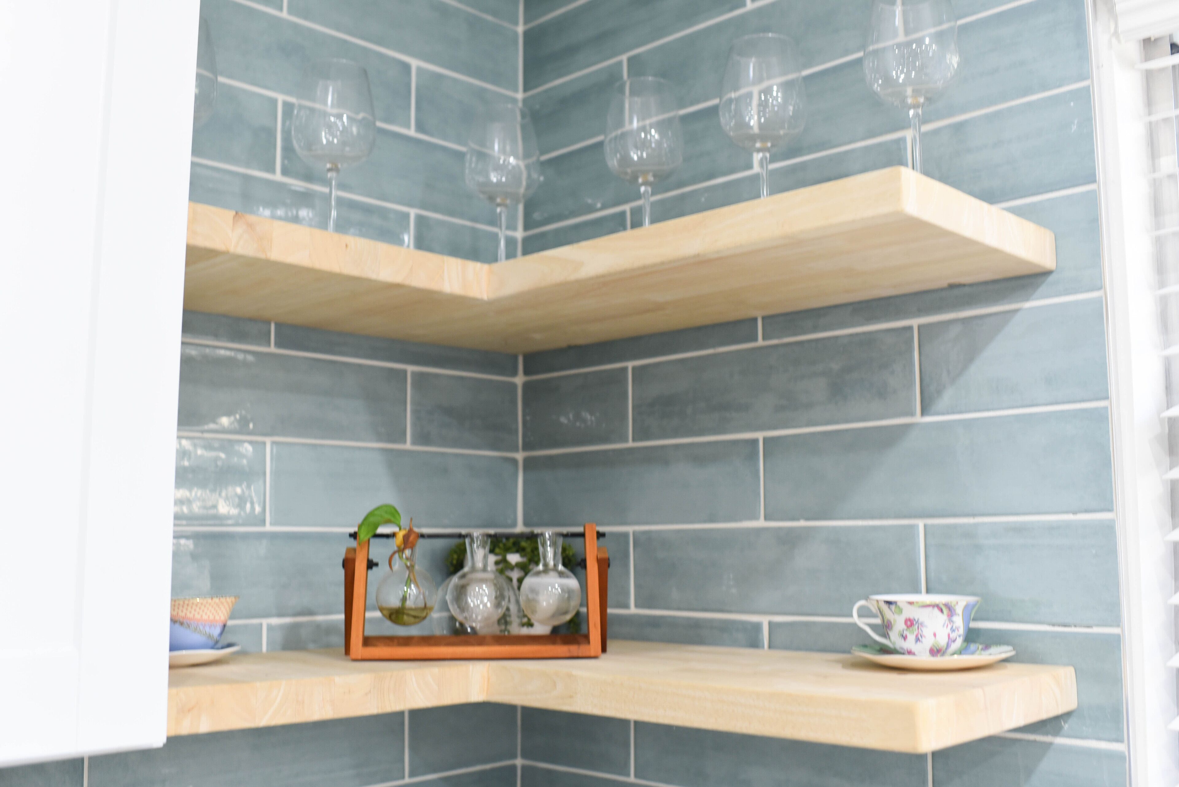 A corner with two wooden shelves against a tiled wall, holding glassware and a floral cup.