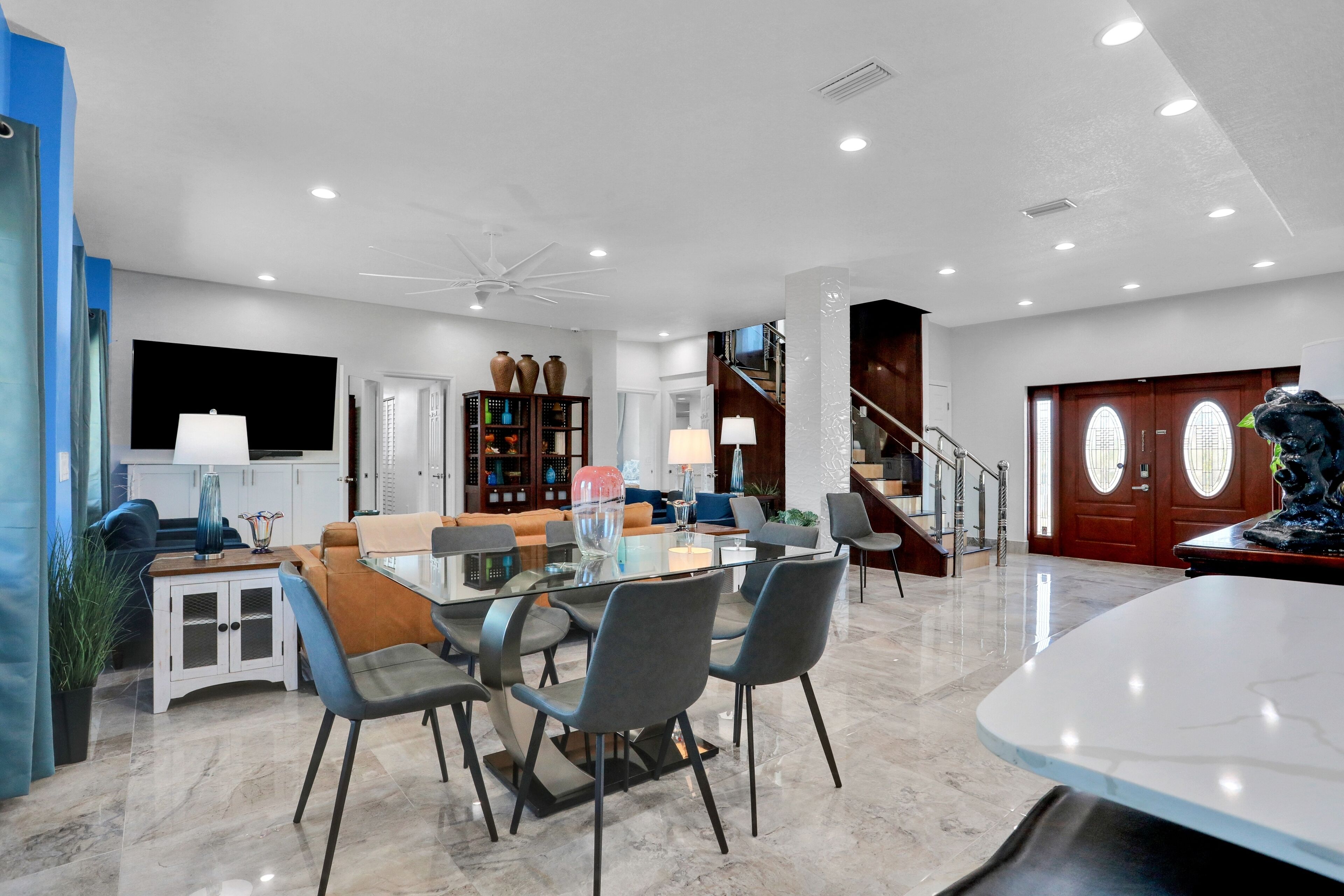 A spacious living area with a glass dining table, comfortable seating, a staircase, and a large wooden door.