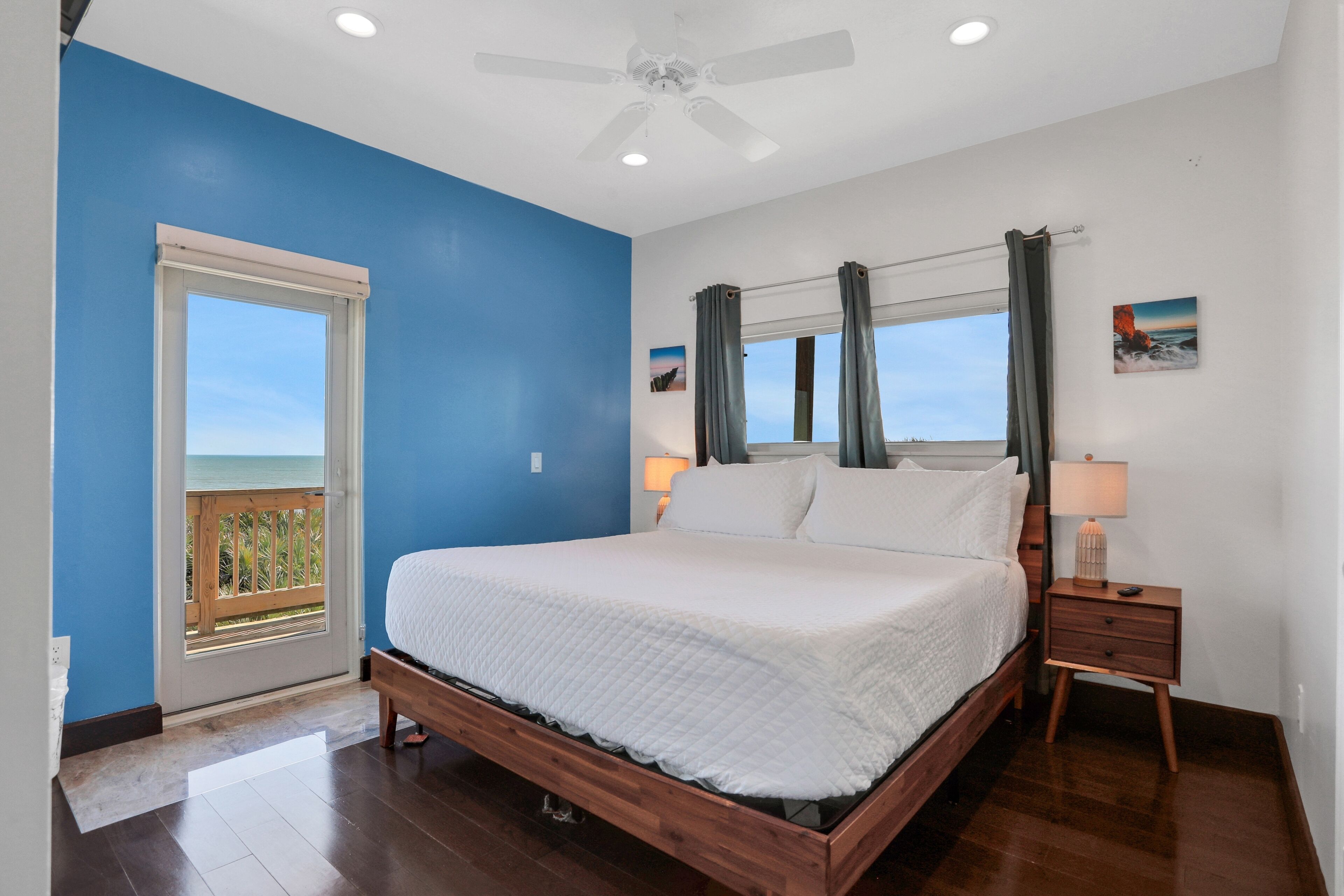 A bedroom with a large bed, a nightstand, a ceiling fan, a window with curtains, and a view of the ocean.