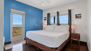 Una camera da letto con un letto grande, un comodino, una ventola a soffitto, una finestra con tende e vista sull'oceano.
