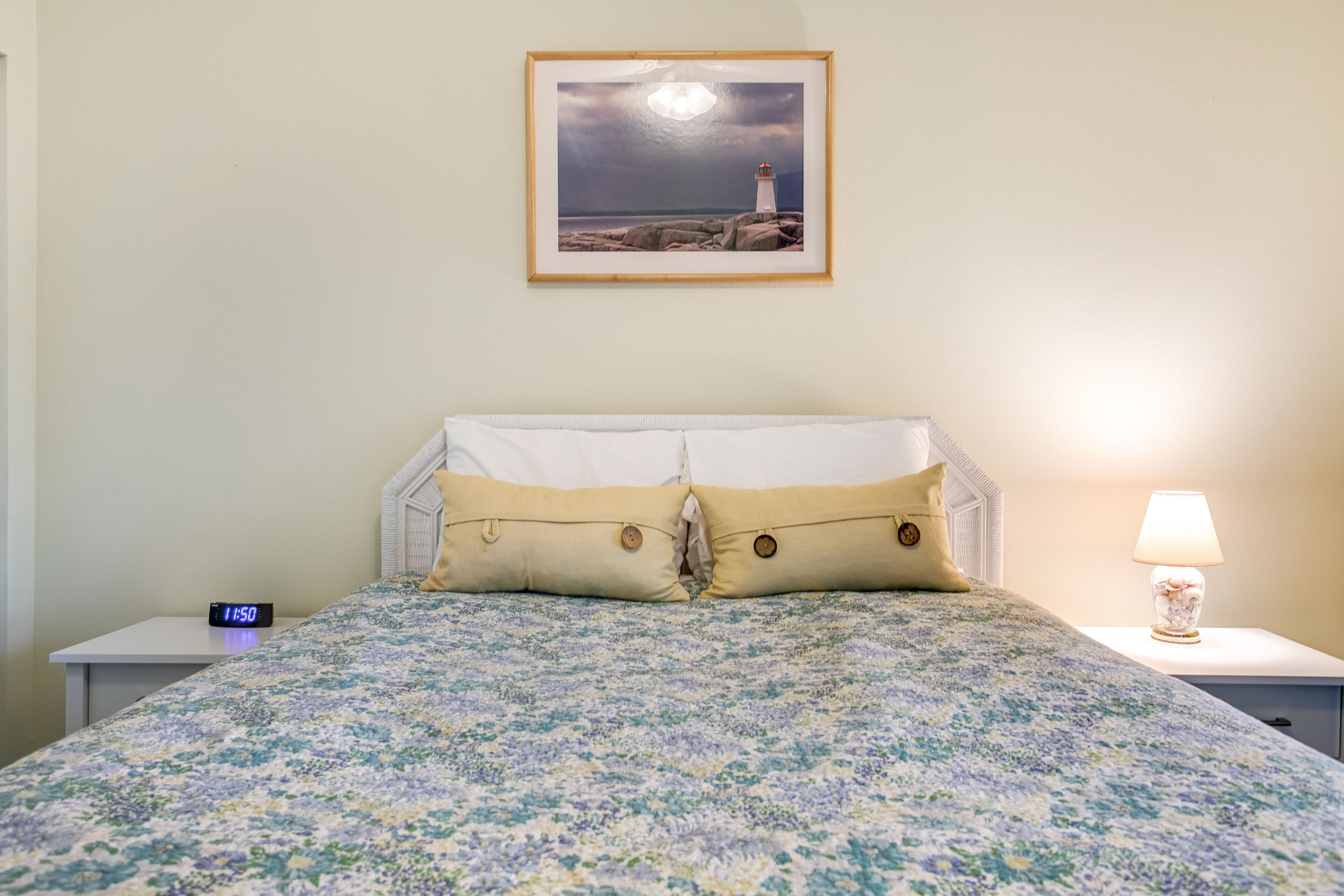 Una cama bien hecha con una colcha estampada con motivos florales, una mesita de noche con una lámpara, un reloj y un cuadro con un faro en la pared.