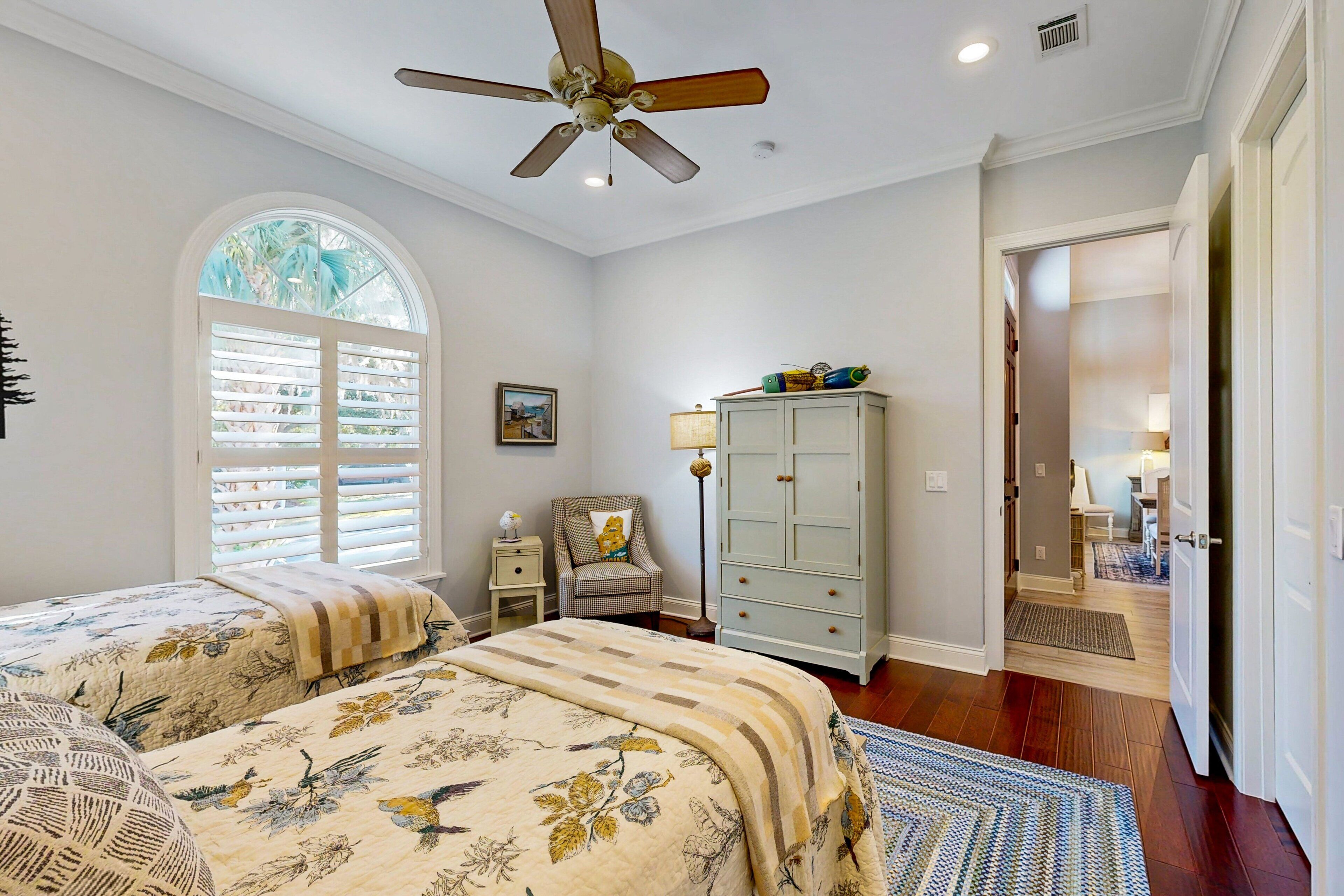 A bedroom with a large bed, a ceiling fan, a window with blinds, a nightstand, a chair, a cabinet, and a door leading to another room.