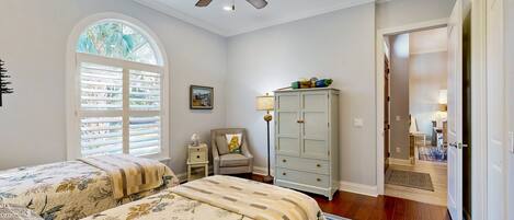 A bedroom with a large bed, a ceiling fan, a window with blinds, a nightstand, a chair, a cabinet, and a door leading to another room.