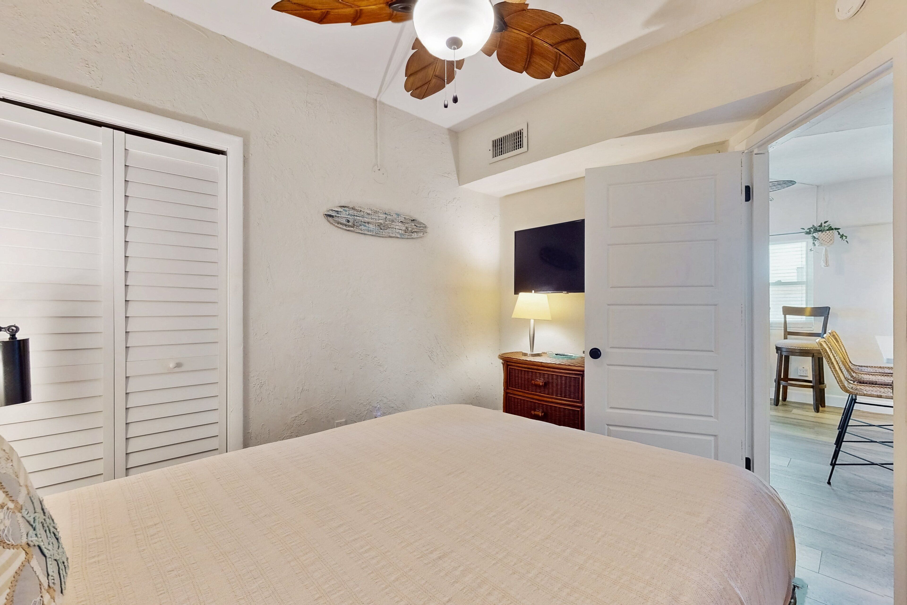 A bedroom with a bed, a ceiling fan, a wall-mounted TV, and a door leading to another room.