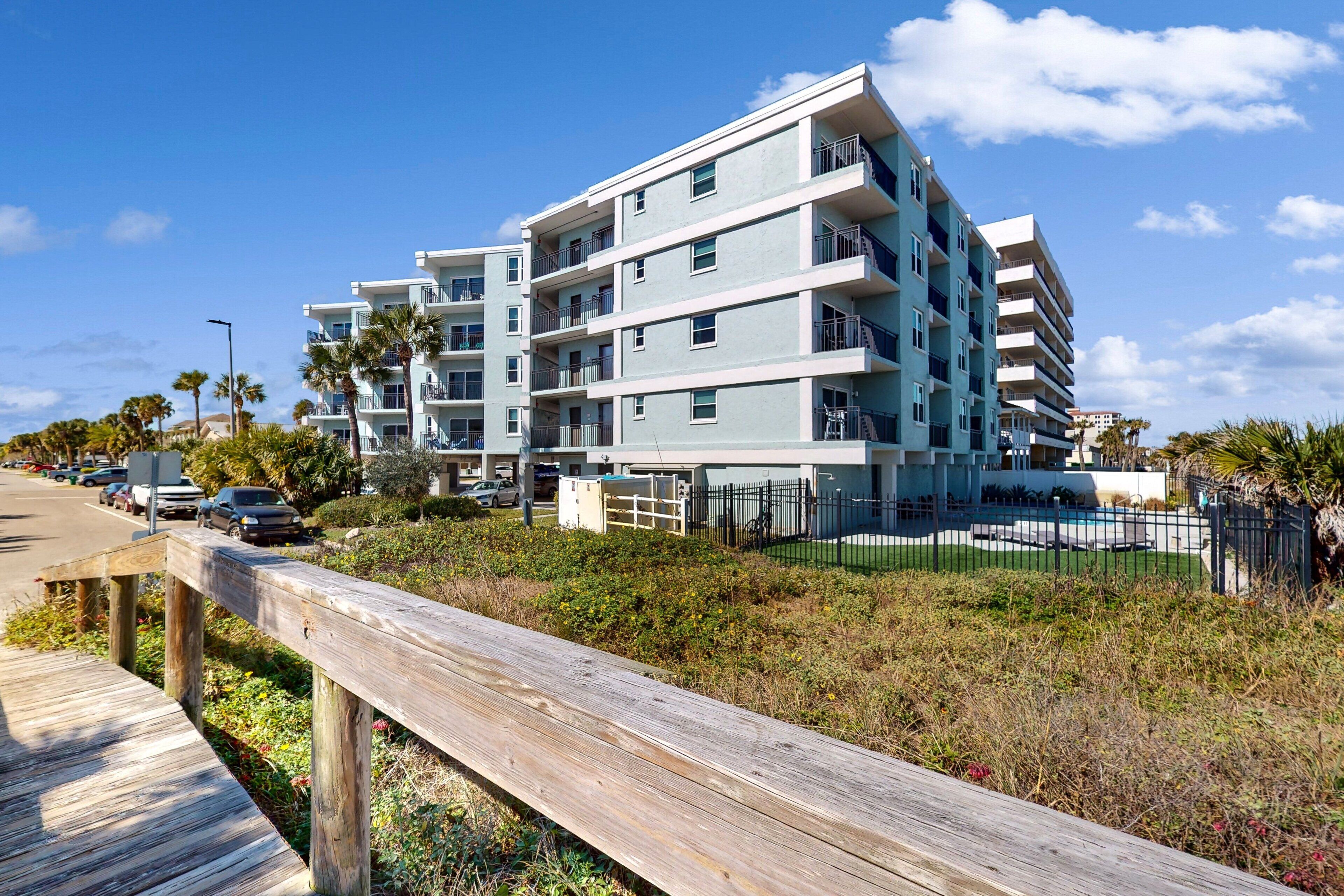 A multi-story residential building with balconies, a wooden boardwalk, and a fenced pool area.