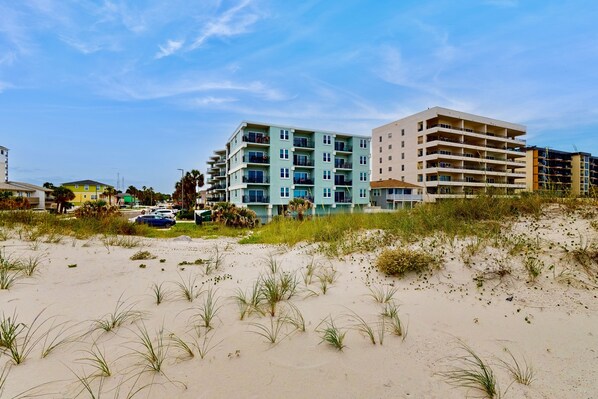 A beachfront hotel with multiple floors, a sandy beach in the foreground, and a clear blue sky. - Modern Oceanfront Condo With Outdoor Pool, Balcony, & Washer/dryer (Jacksonville Beach)