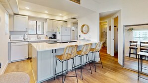 A modern kitchen with white cabinets, a central island with bar stools, and a 'Welcome' sign. - Modern Oceanfront Condo With Outdoor Pool, Balcony, & Washer/dryer (Jacksonville Beach)