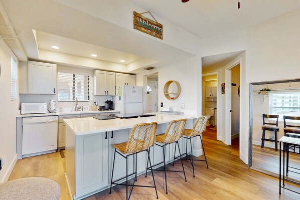 A modern kitchen with white cabinets, a central island with bar stools, and a 'Welcome' sign. - Modern Oceanfront Condo With Outdoor Pool, Balcony, & Washer/dryer (Jacksonville Beach)
