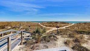 Ein hölzerner Steg führt zu einem Strand mit Dünen und wolkenlosem Himmel.