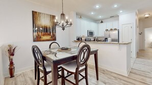 A dining area with a glass table, wooden chairs, and a chandelier. There is a kitchen in the background with white cabinets and appliances. - Immaculate Townhome in Upscale Community w/ Pool & Balcony - Near Beach (Palm Coast)
