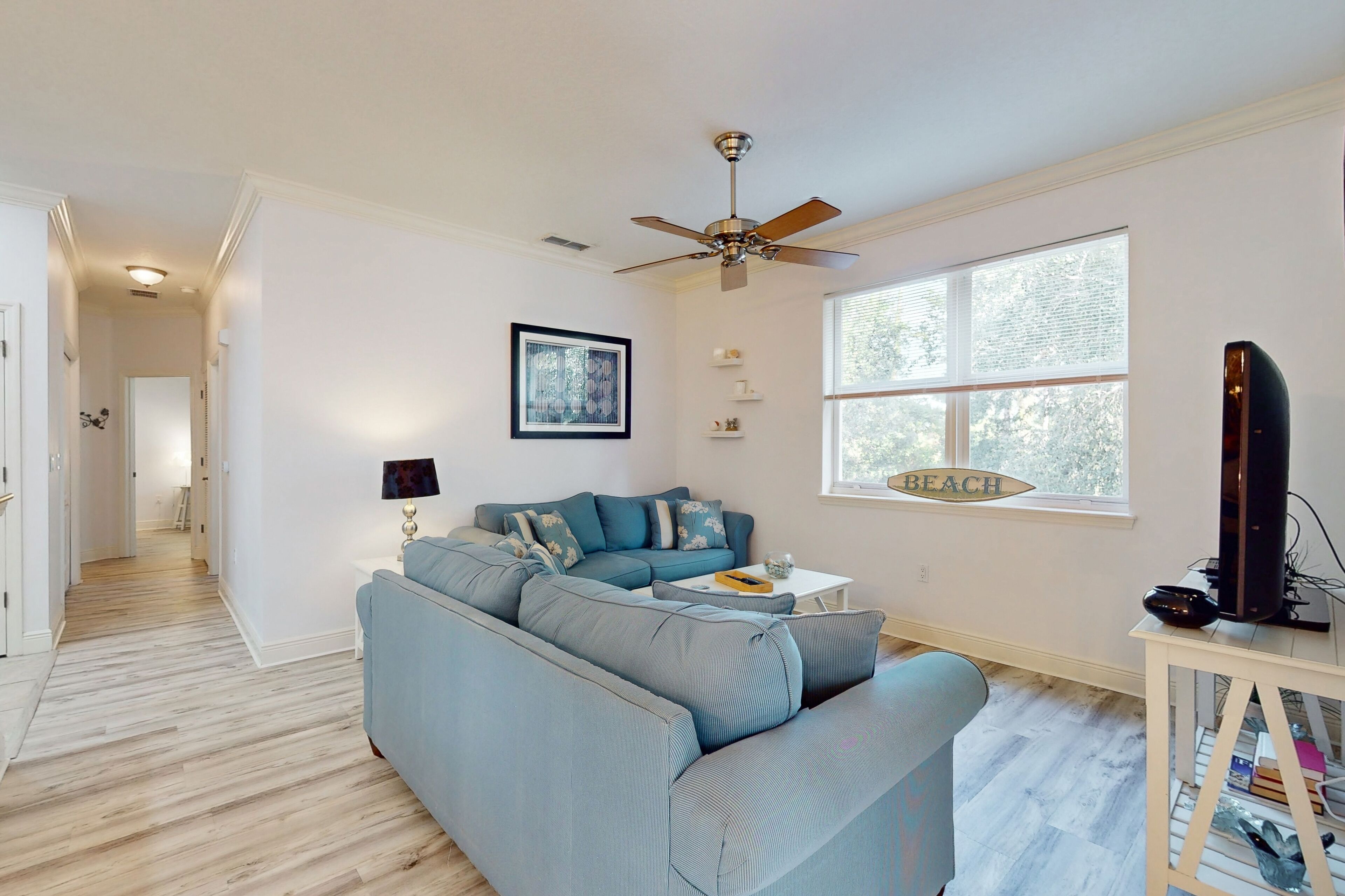 A living room with a sectional sofa, a ceiling fan, a large window, and a TV on a stand.