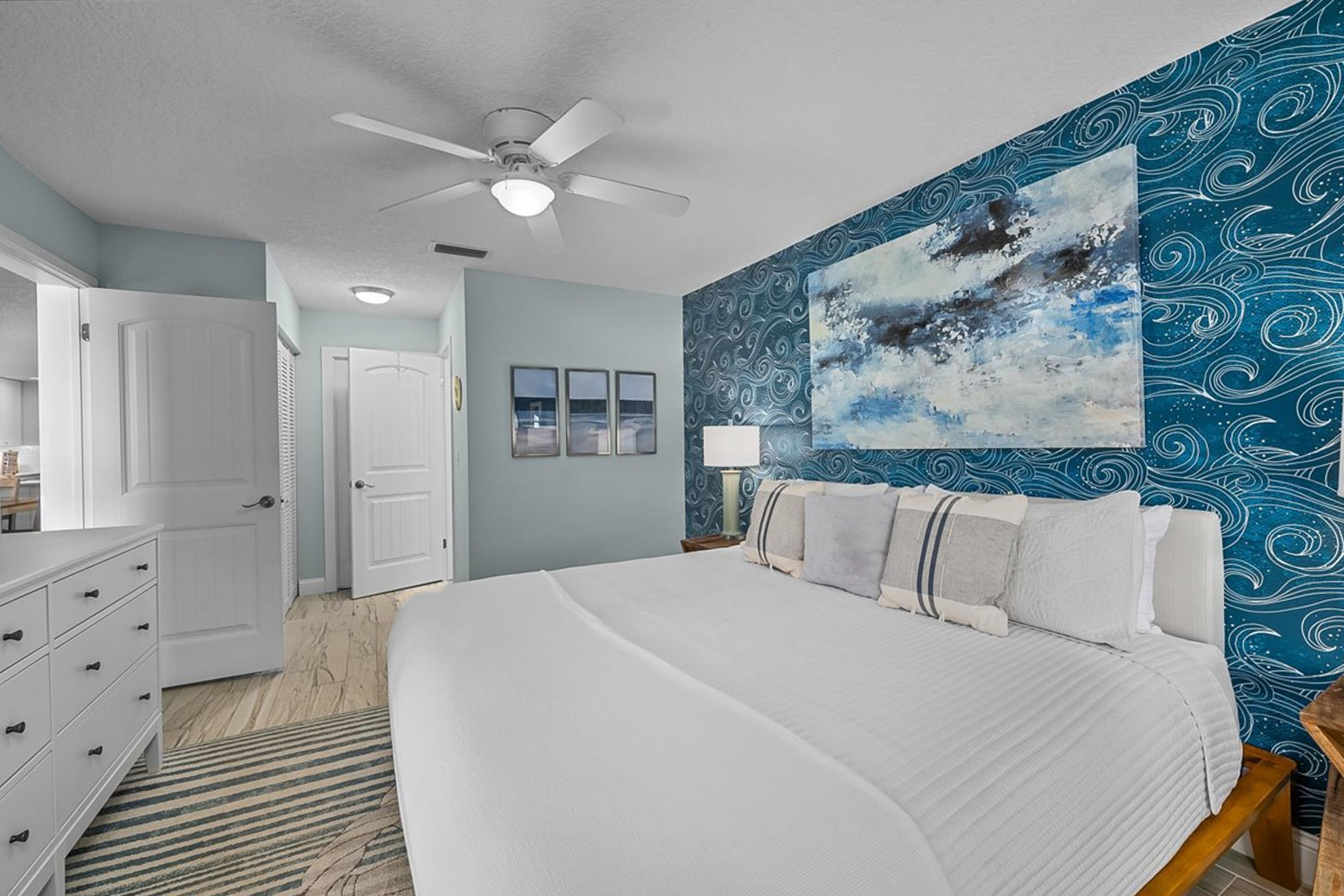 A bedroom with a large bed, a ceiling fan, a blue patterned wall, and a door leading to another room.
