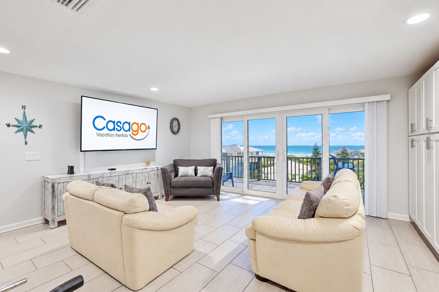 A spacious living room with a large TV screen, two sofas, and a balcony with a view of the sea.