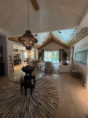 A spacious living area with a high ceiling, a large chandelier, and a view through a sliding door to the outdoors. - Lakefront Home With Private Pool & Outdoor Space for Entertaining (St. Simons Island)