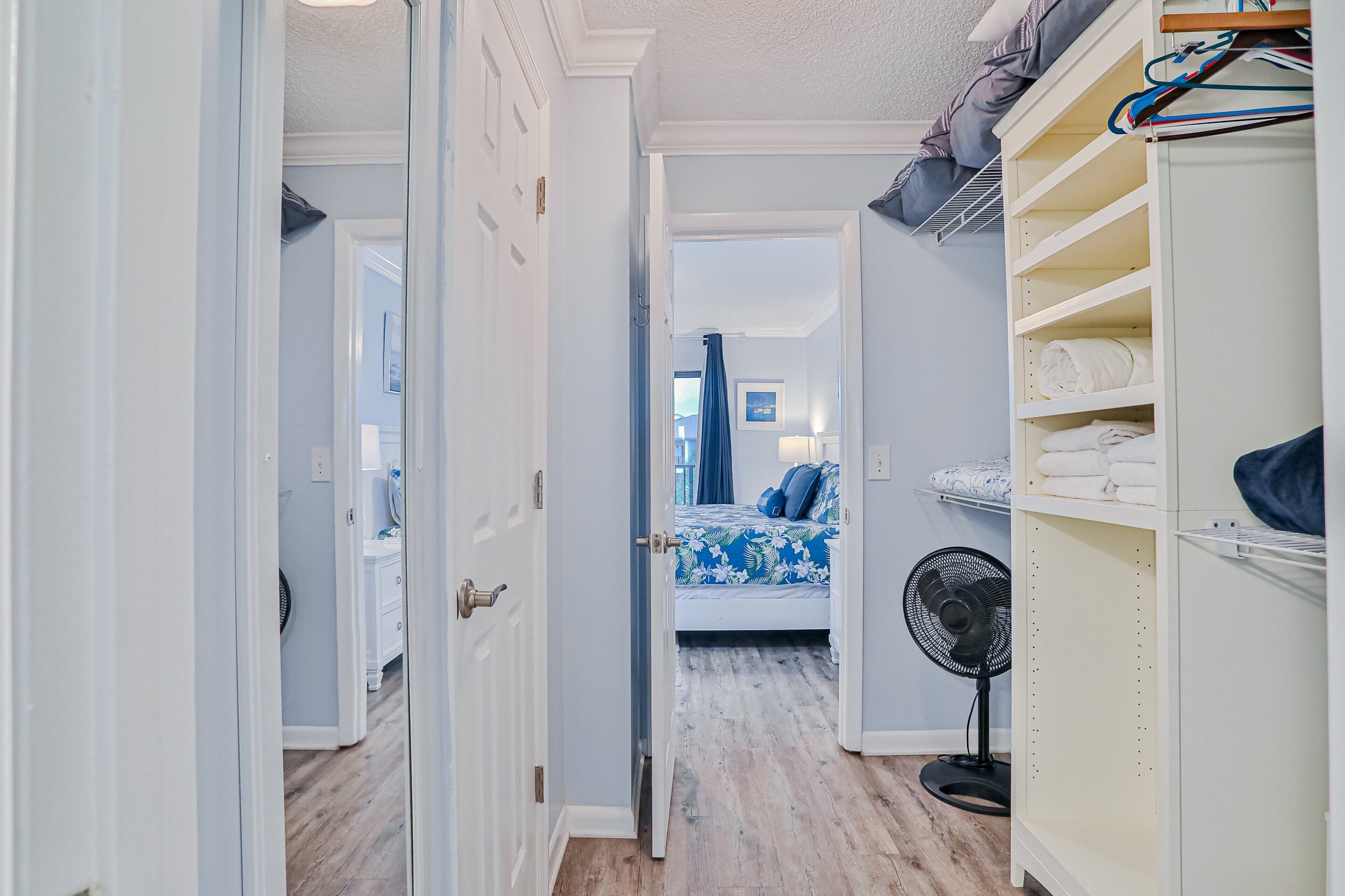 A narrow hallway leading to a bedroom with a bed, a fan, and a closet with shelves.