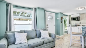 A living room with a grey sofa, a dining area with a white chair, and a kitchen with white cabinets and a microwave. - Home 1 Block From 2 Beaches W/private Pool, High-speed Wifi & Central AC (St. Augustine)