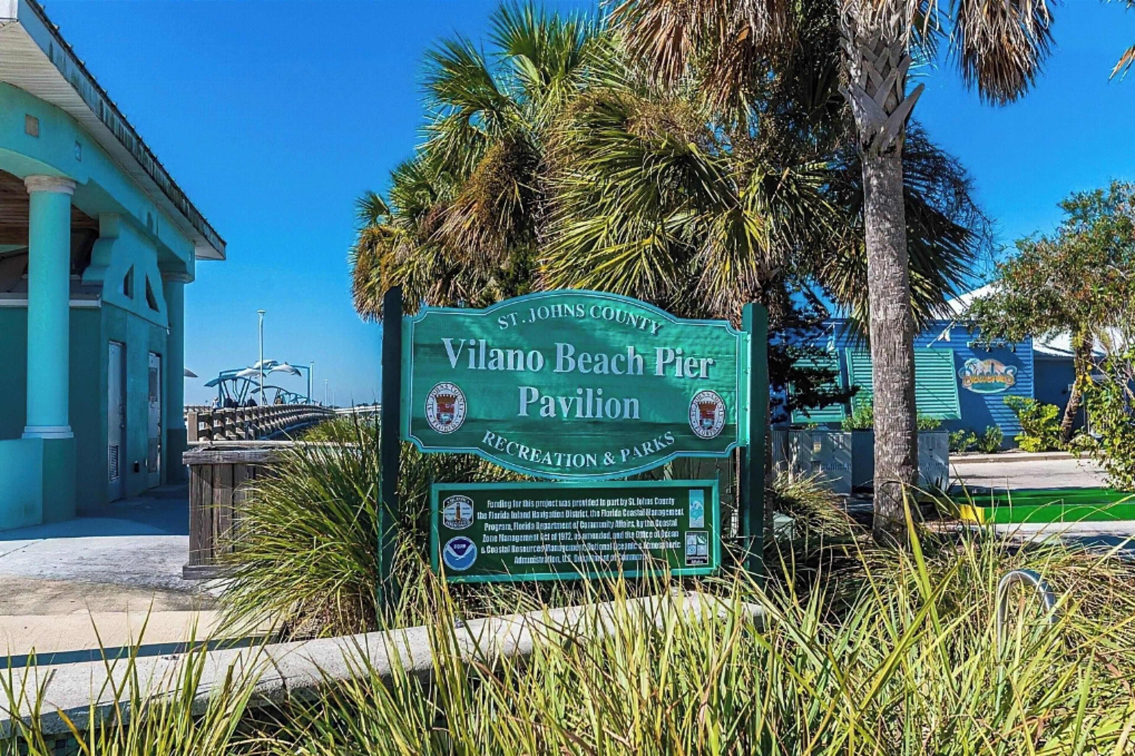 A sign for Vilano Beach Pier Pavilion in St. Johns County, Florida, with a clear blue sky and palm trees in the background.