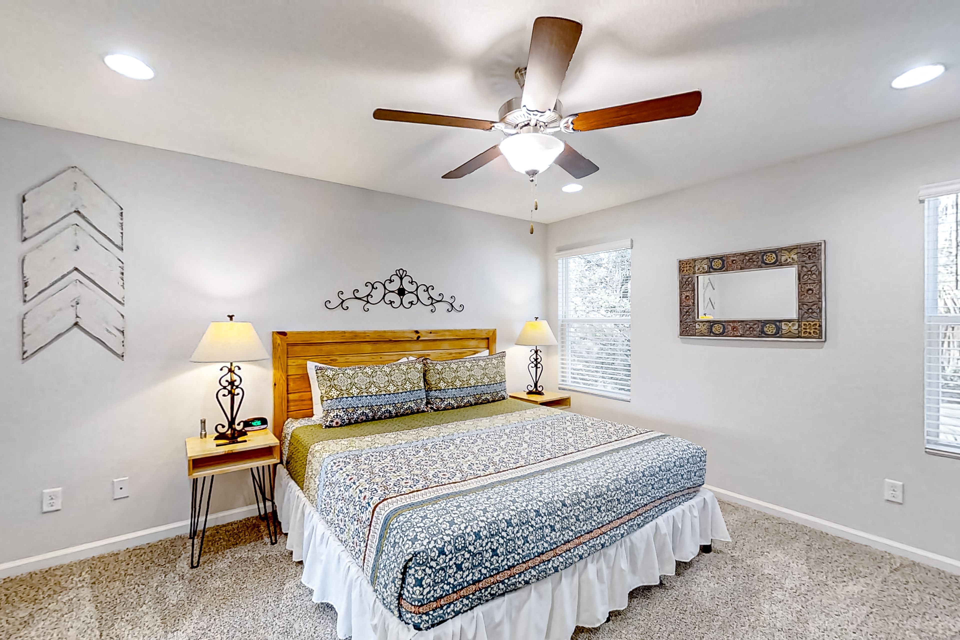 Une chambre à coucher comprenant un lit, deux tables de chevet, un ventilateur de plafond, une fenêtre avec des stores et des œuvres d’art décoratives au mur.