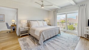 Une chambre avec un grand lit, un ventilateur de plafond, une table de chevet avec une lampe, un bureau avec une chaise et une vue sur l’extérieur grâce à une grande fenêtre.