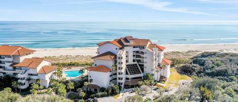 A multi-story hotel with a pool, beachfront view, and a parking area.