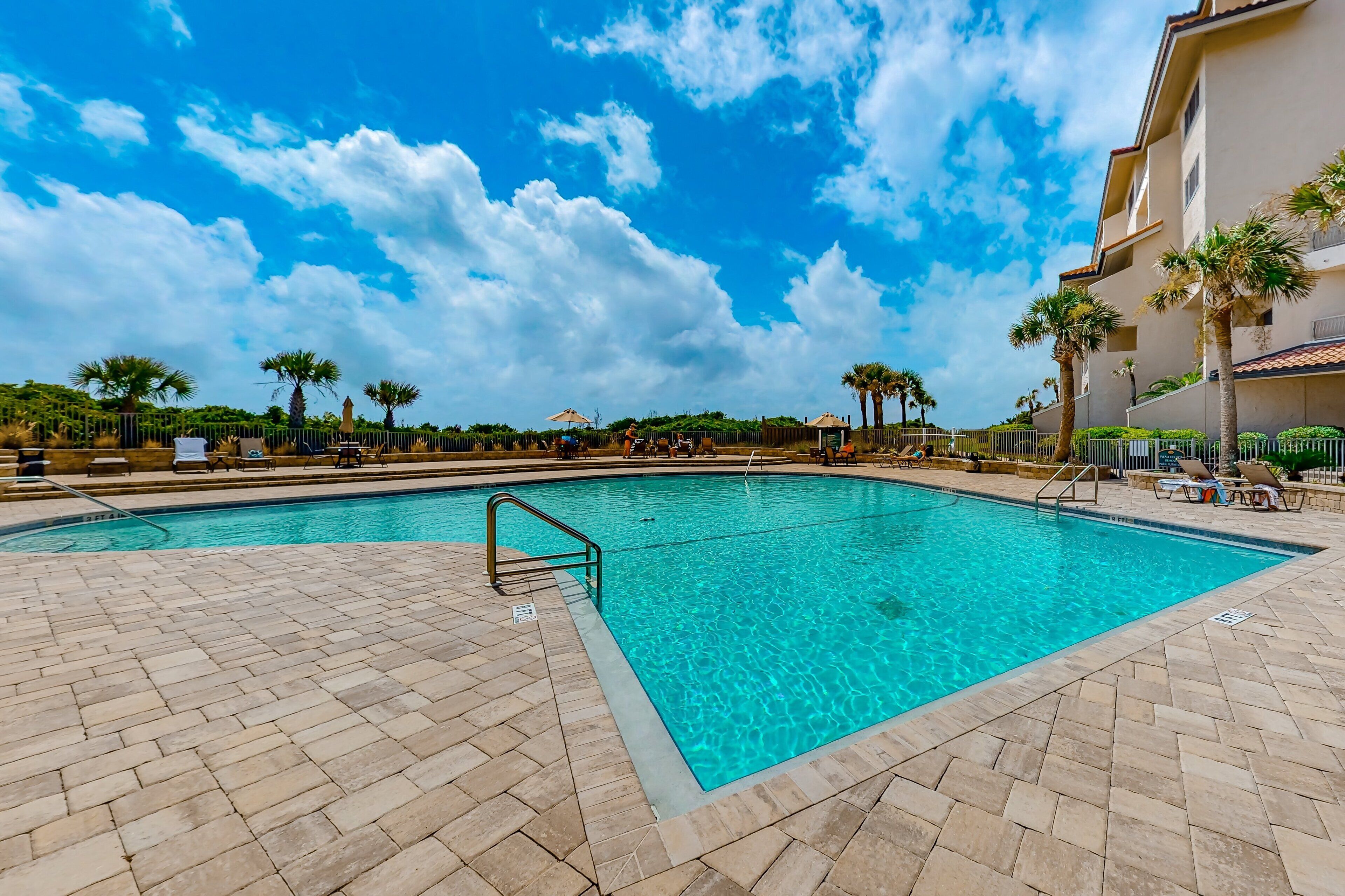 A swimming pool with clear blue water, surrounded by a tiled deck and palm trees.