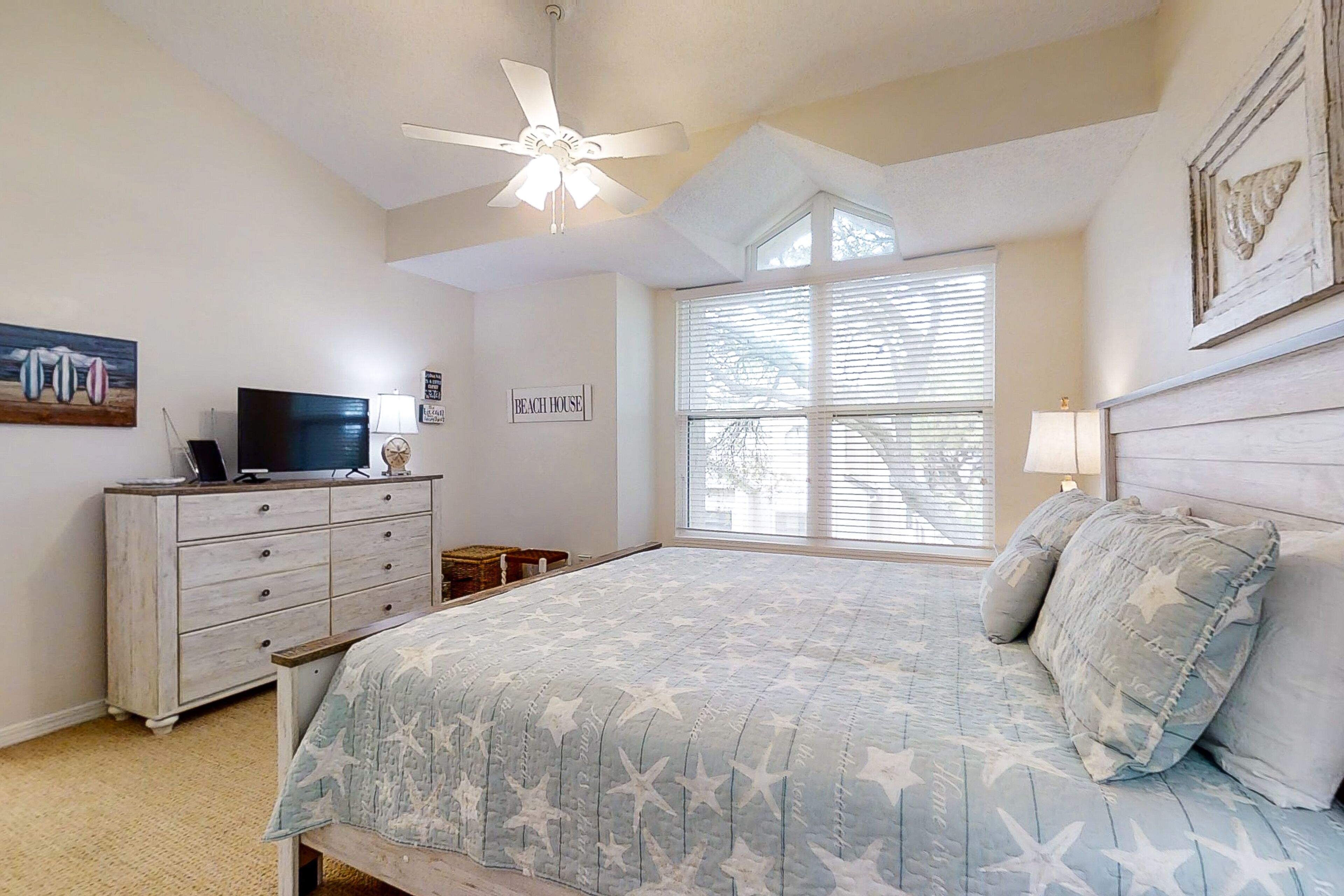 A bedroom with a bed, a ceiling fan, a TV on a dresser, and a window with blinds.