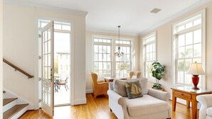 A spacious living room with hardwood floors, a sofa, chairs, a chandelier, and large windows. - Luxury Cottage With Pools & Multiple Decks, 1/2 Block to Beach (St. Simons Island)