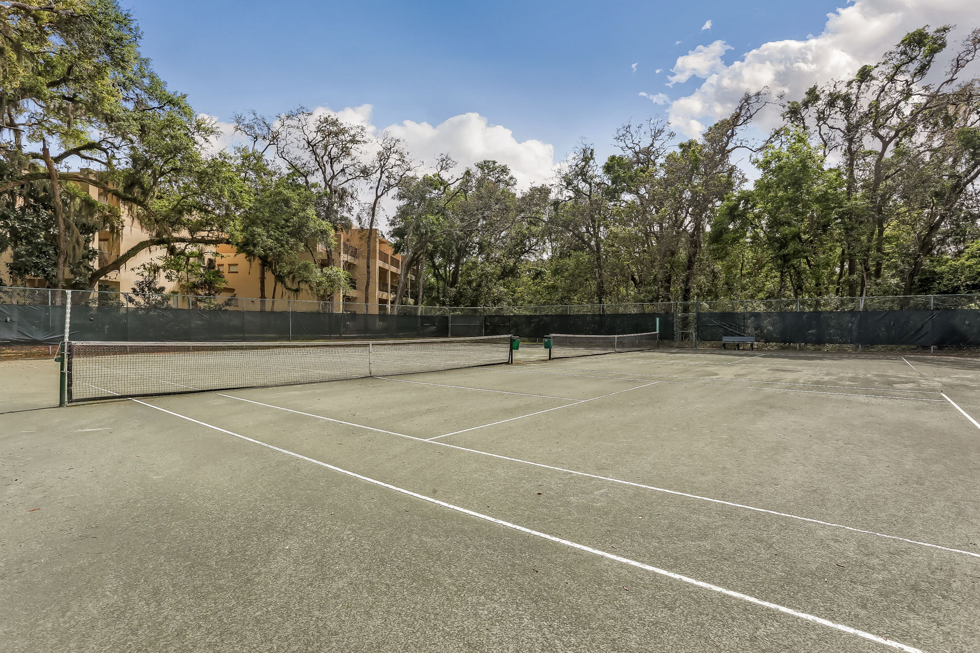 An outdoor tennis court with a net, surrounded by trees and a building in the background.