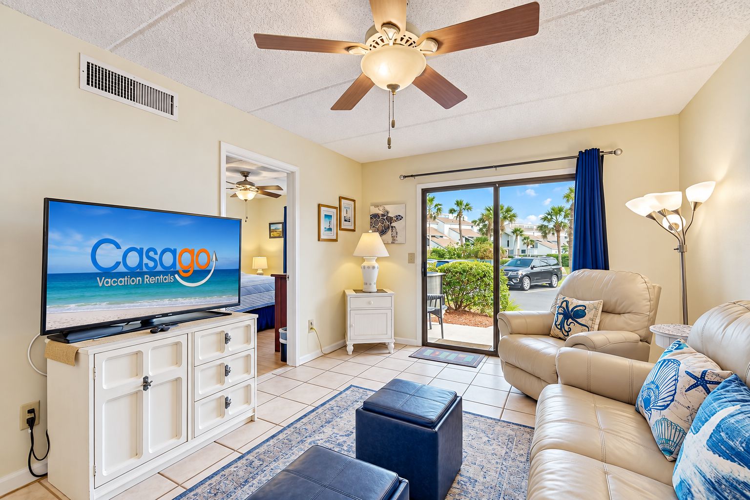 A hotel room with a television displaying 'Casalgo Vacation Rentals', a ceiling fan, a sofa, a coffee table, a lamp, and a view of palm trees through the door.