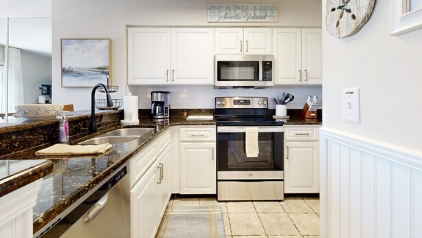 A modern kitchen with white cabinetry, a granite countertop, stainless steel appliances, and a beach-themed sign. - Newly Decorated, Oceanfront Townhouse With Patio, Wifi, W/D & Shared Pool (Fernandina Beach)