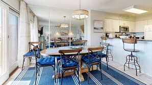 A dining area with a round table and chairs, a kitchen area with a microwave and refrigerator, and a large window with curtains. - Newly Decorated, Oceanfront Townhouse With Patio, Wifi, W/D & Shared Pool (Fernandina Beach)