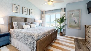 A bedroom with a large bed, a ceiling fan, framed botanical prints, a potted plant, and a beach view through the window. - Newly Decorated, Oceanfront Townhouse With Patio, Wifi, W/D & Shared Pool (Fernandina Beach)