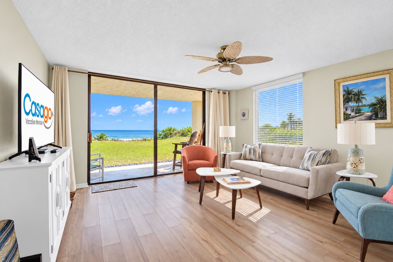 A spacious living room with a large sliding glass door leading to a balcony, a ceiling fan, a flat-screen TV, and a view of the ocean.
