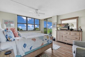 Une chambre à coucher avec un lit, un ventilateur de plafond, une commode en bois et une grande fenêtre donnant sur l’océan.