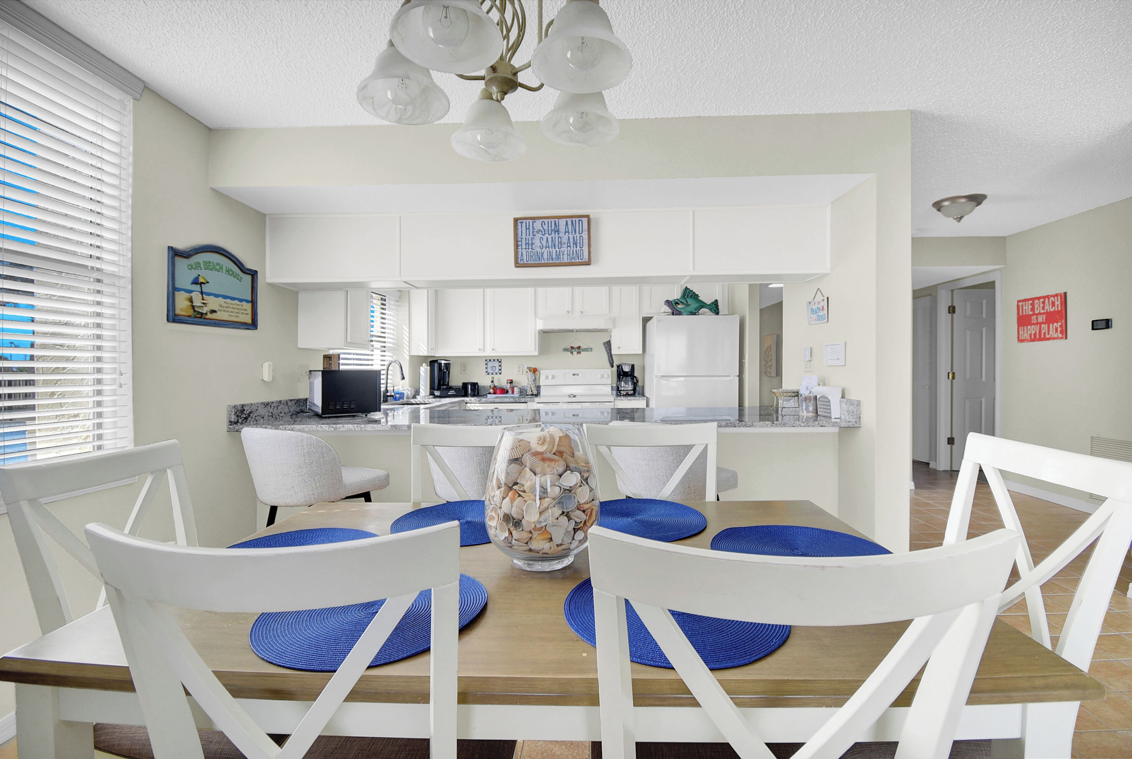 A dining area with a wooden table, white chairs, and blue placemats. A glass vase with seashells is on the table. The kitchen is visible in the background with white cabinets and a microwave.