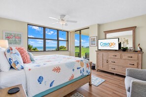 Un dormitorio con cama, ventilador de techo, ventana grande con vistas, televisor y cómoda de madera.