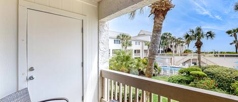 A balcony with a wicker chair and a wooden bench, overlooking a pool and palm trees.