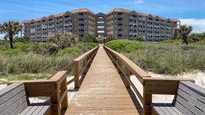 A wooden boardwalk leading to a multi-story hotel with balconies, surrounded by greenery and sand. - Ocean View Condo With Pool Access, Wifi, & Central AC - Snowbirds OK (Palm Coast)