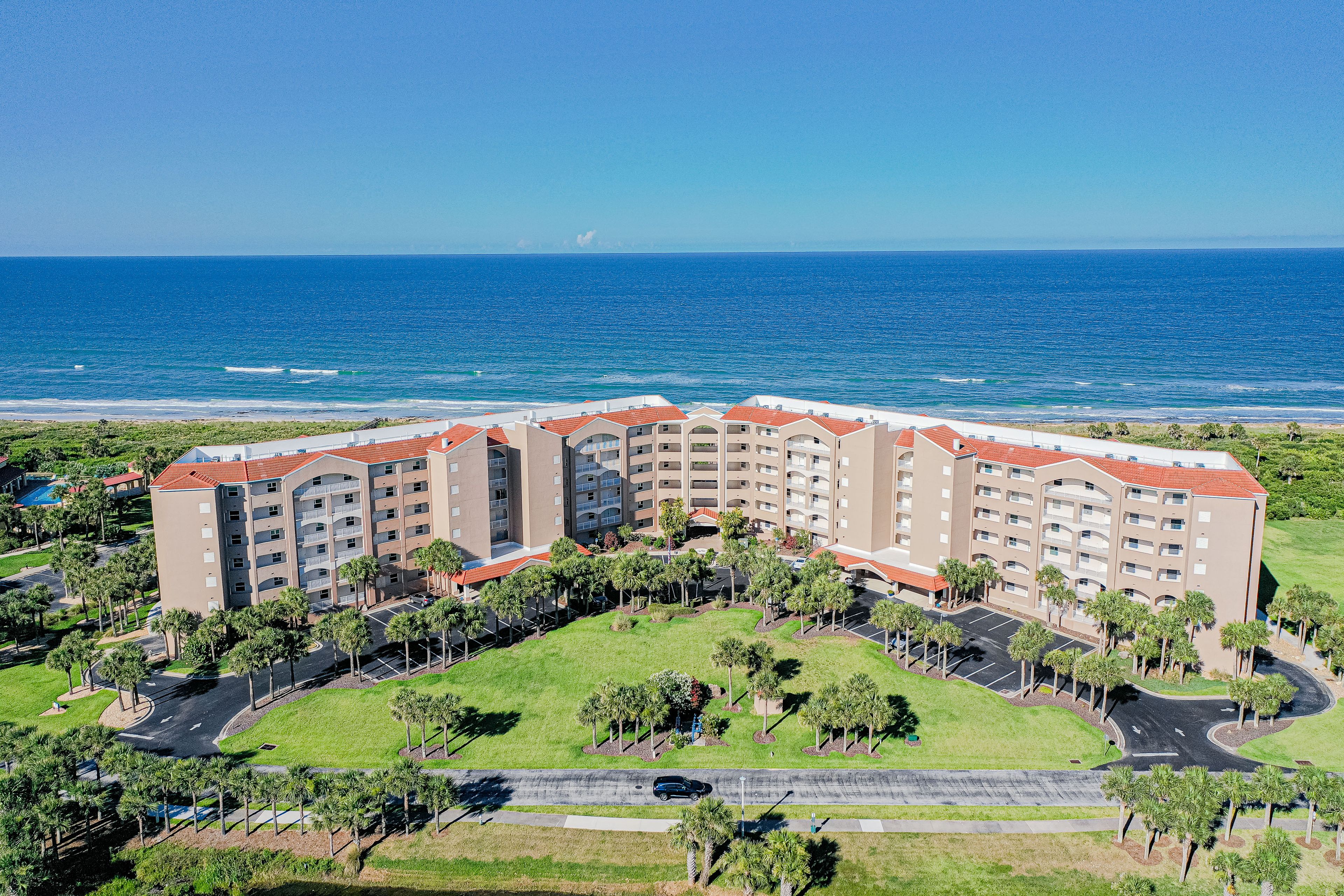 A multi-story hotel with a curved roof, surrounded by a landscaped area with palm trees and a parking lot.