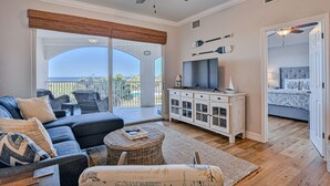 A spacious living room with a large sofa, a television, and a balcony with a view. - Ocean View Condo With Pool Access, Wifi, & Central AC - Snowbirds OK (Palm Coast)