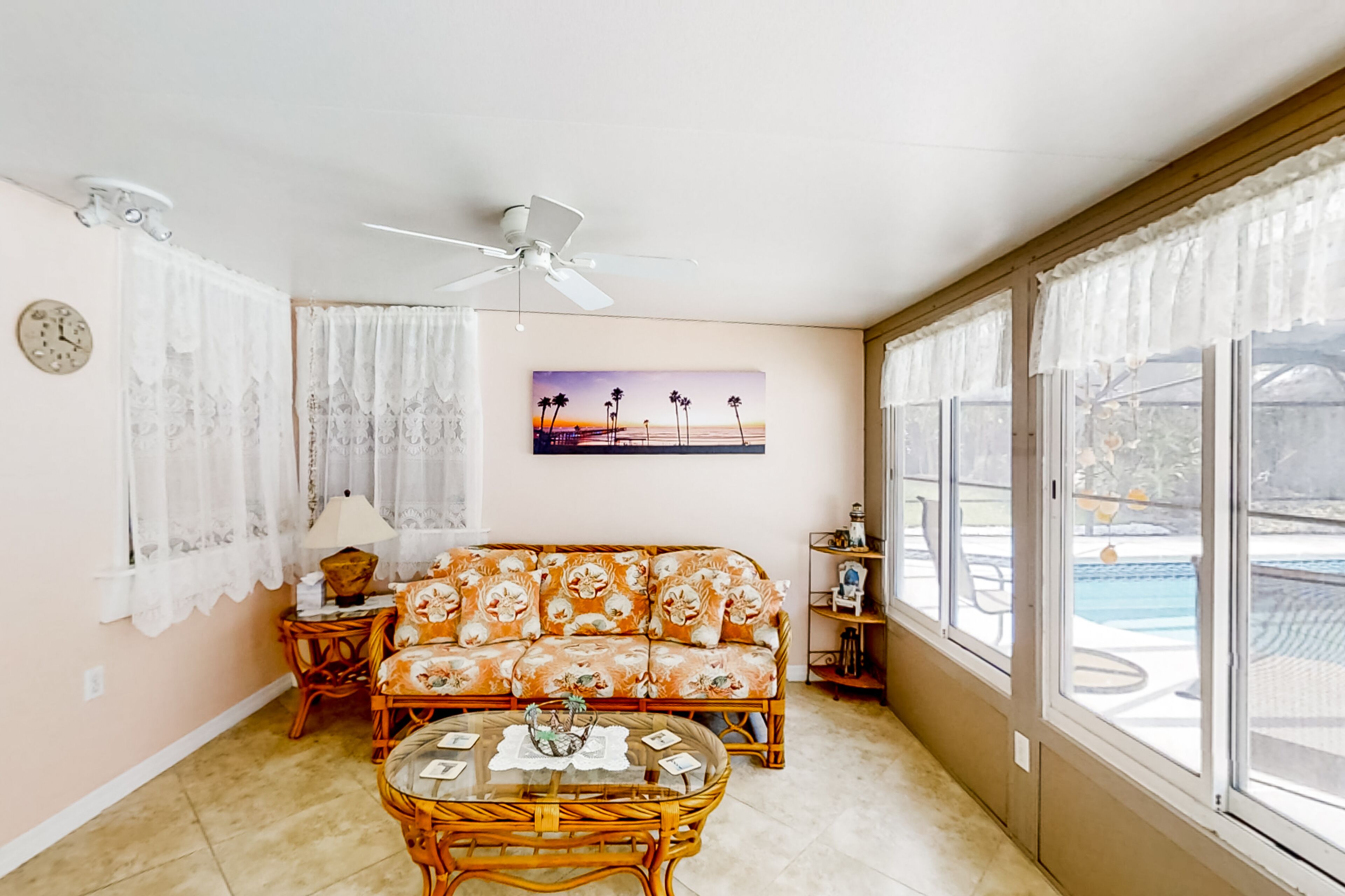 A living room with a sofa, a coffee table, a ceiling fan, lace curtains, and a painting of a sunset.
