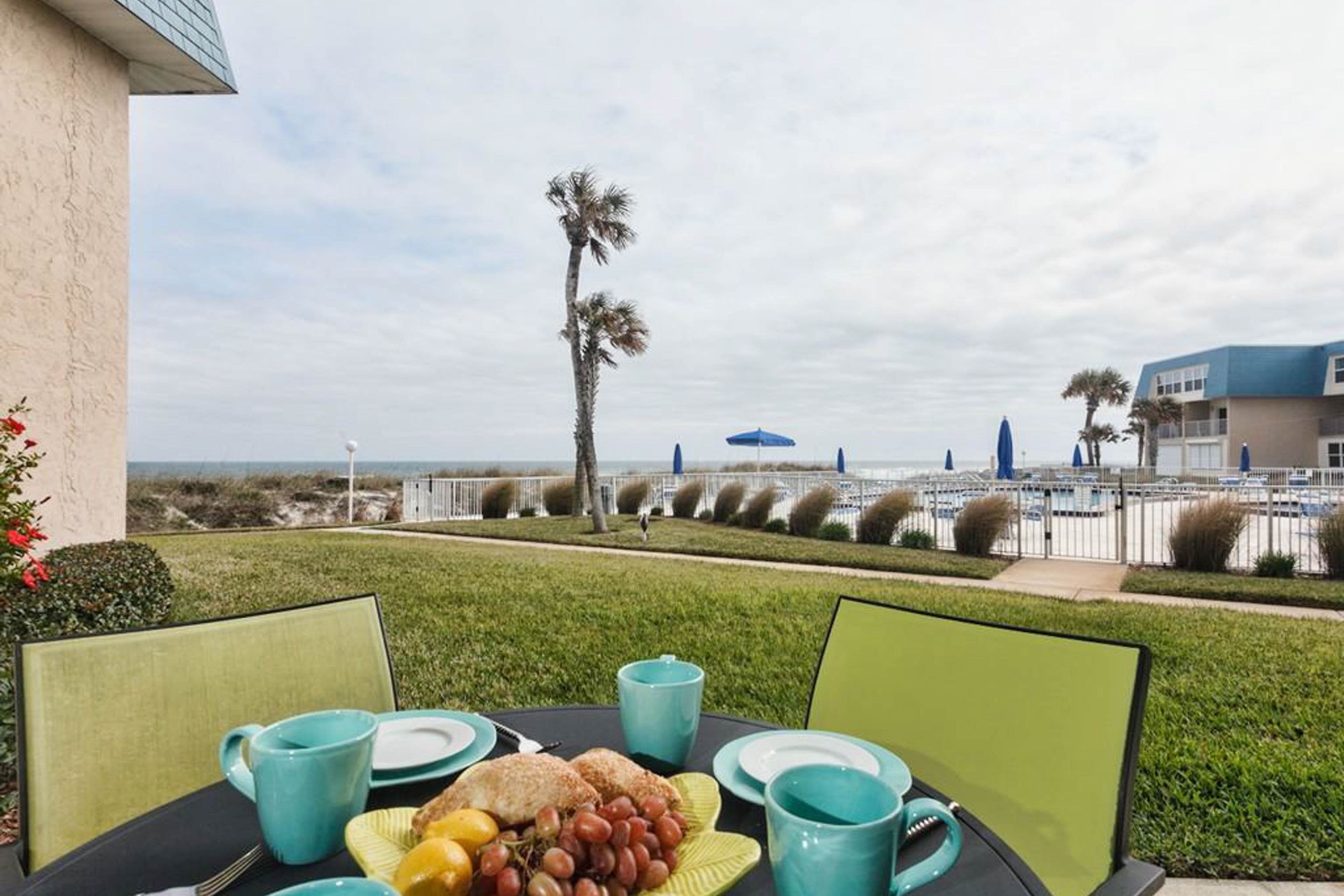 Taula preparada amb articles per esmorzar, com fruita i pastissos, amb vista a una platja amb palmeres i una tanca.