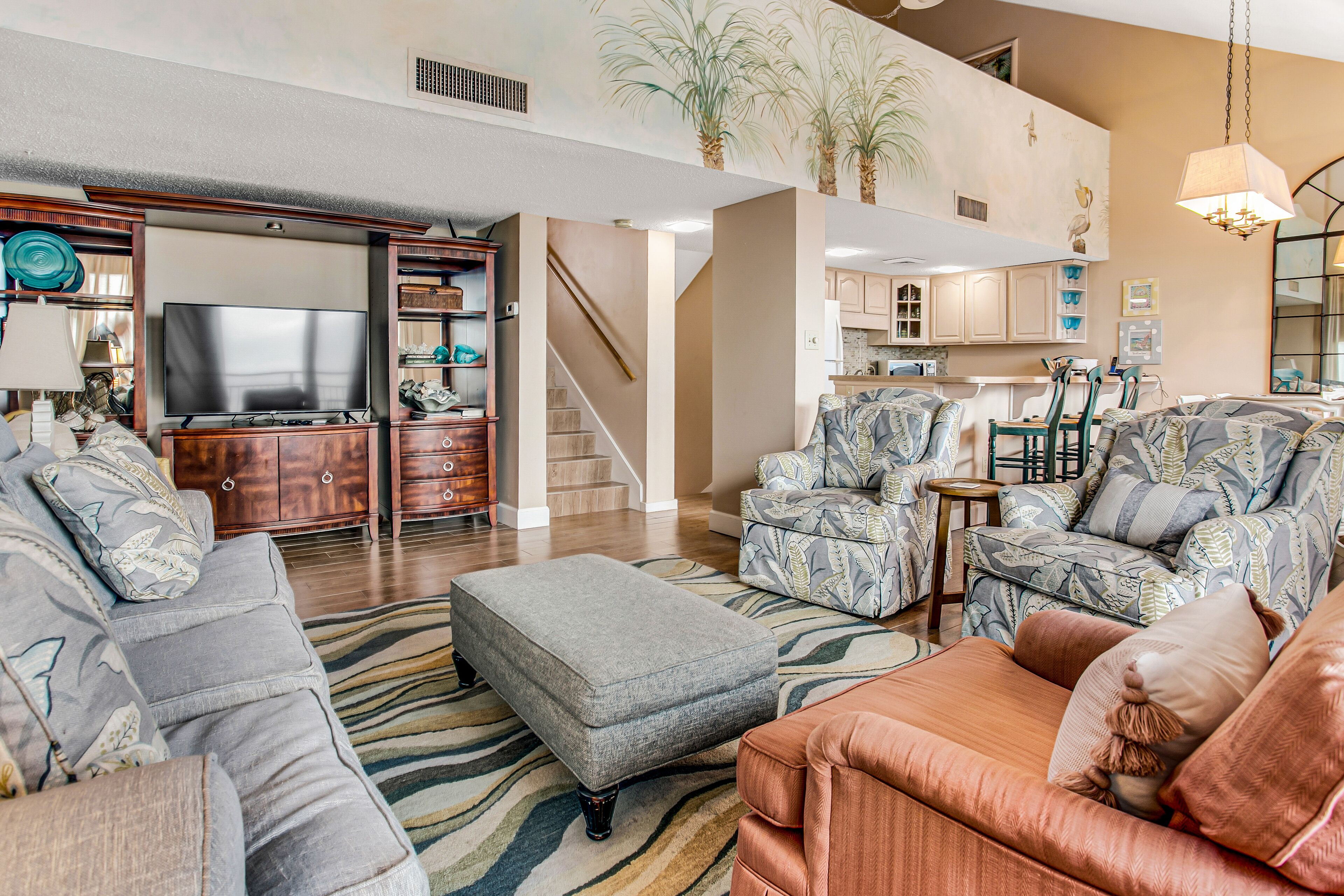 A spacious living room with a large TV, patterned sofas, and a staircase leading to an upper level.