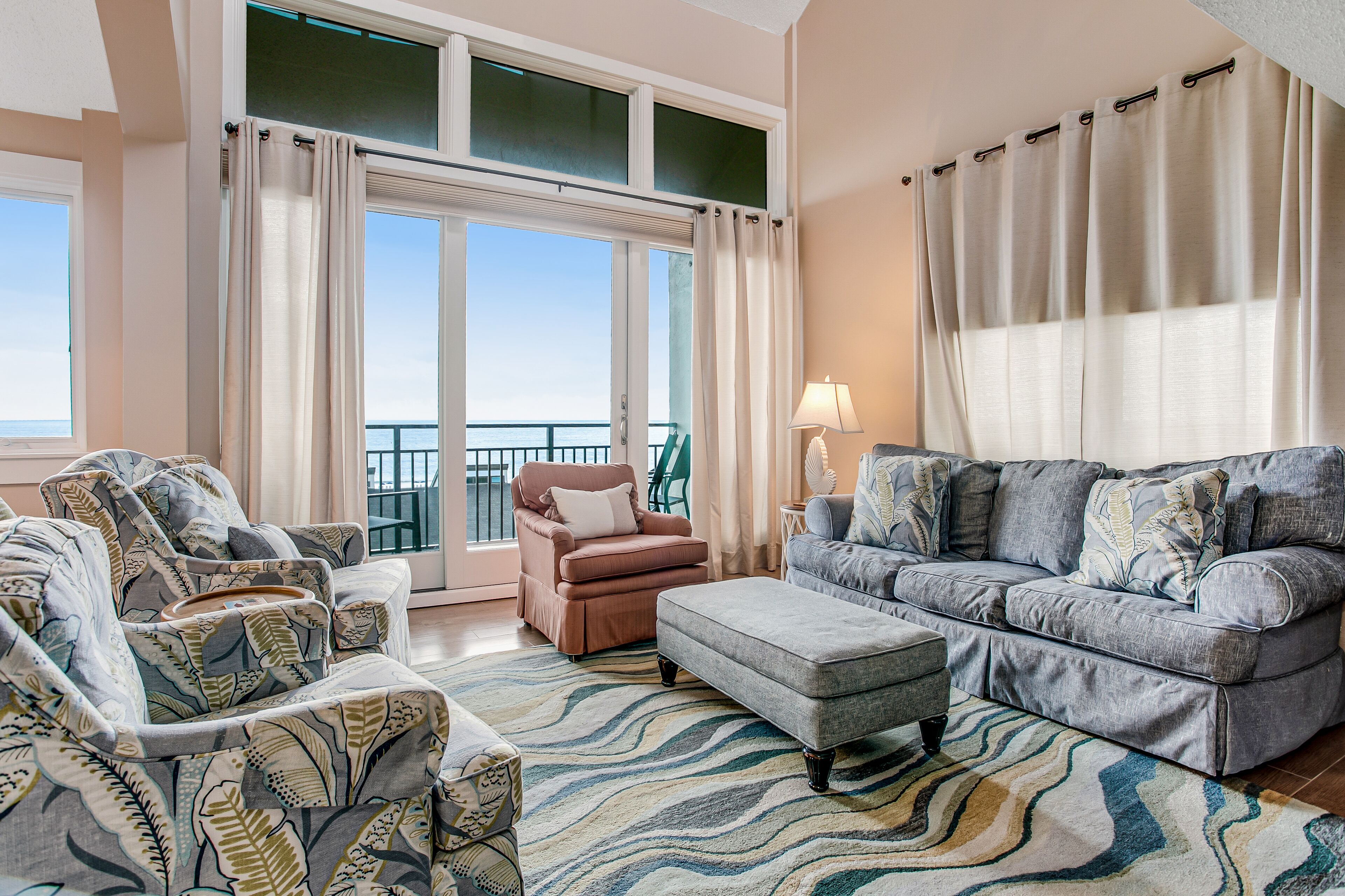 A living room with a sofa, armchair, and ottoman, a patterned rug, and a view of the outdoors through large windows.