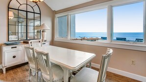 Een eetruimte met een witte tafel en stoelen, een grote spiegel en uitzicht op de oceaan door grote ramen.