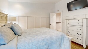 Une chambre à coucher comprenant un lit, une commode, un téléviseur fixé au mur et une porte donnant sur une autre pièce.