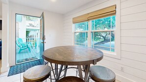 Une pièce avec une table ronde en bois et deux tabourets, une fenêtre avec des stores et une vue sur un balcon équipé de chaises.