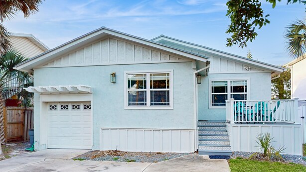 Une maison d’un seul étage, de couleur bleu clair, avec un garage, une véranda et un petit jardin devant, où se trouve un palmier.