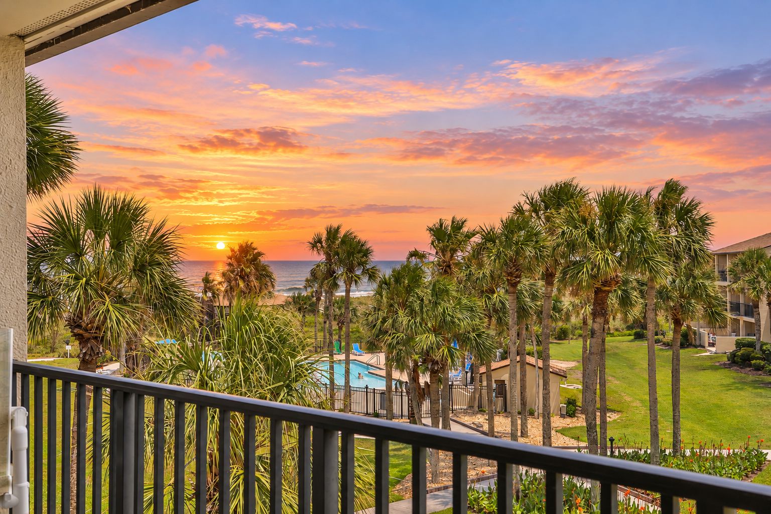 A balcony with a view of a sunset over the ocean, palm trees, and a swimming pool.